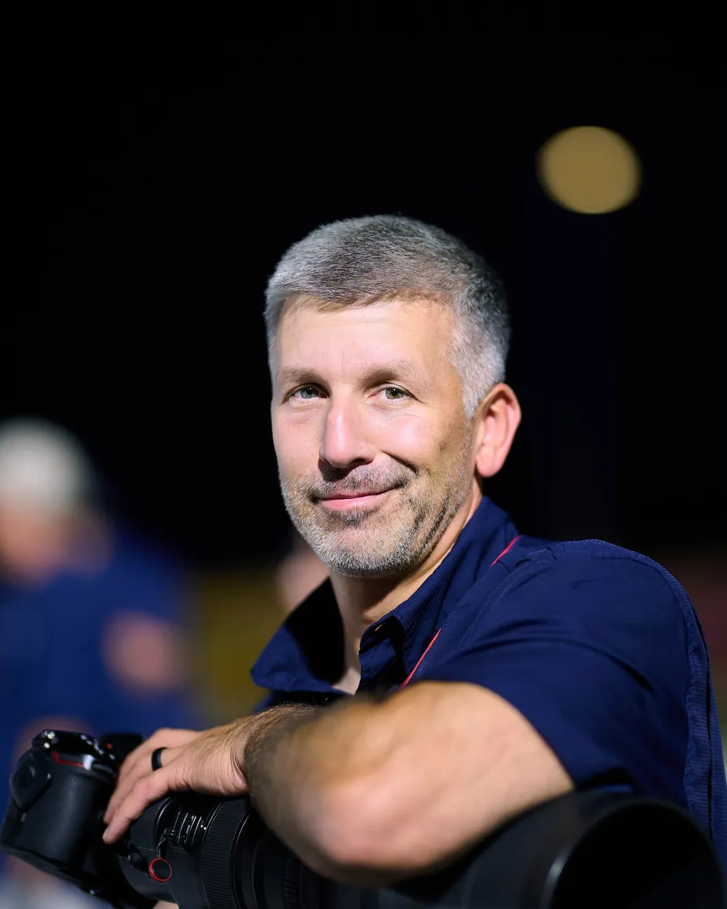 Brian Durkin on the sideline at night, camera and long lens resting on his arm, stadium lights behind him.