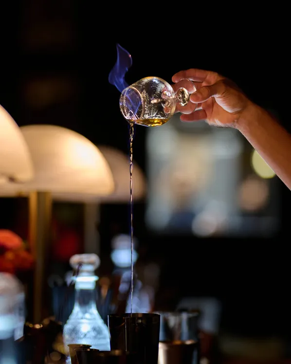 A flaming cocktail poured in a long stream at a dimly lit event bar