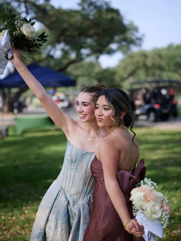 Two friends in formal gowns at an outdoor send-off, one raising her bouquet