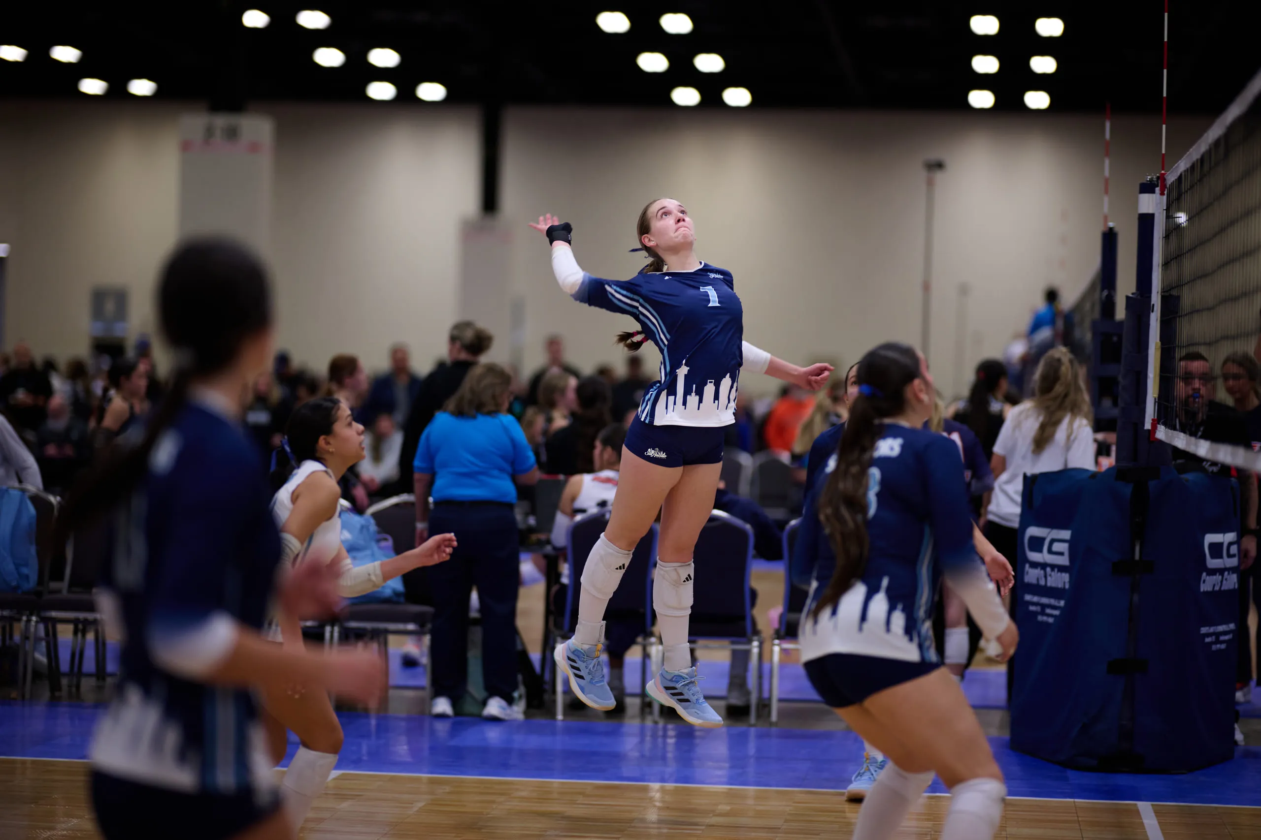 A volleyball player at the top of her jump, arm cocked to hit, teammates blurred around her on a tournament court.