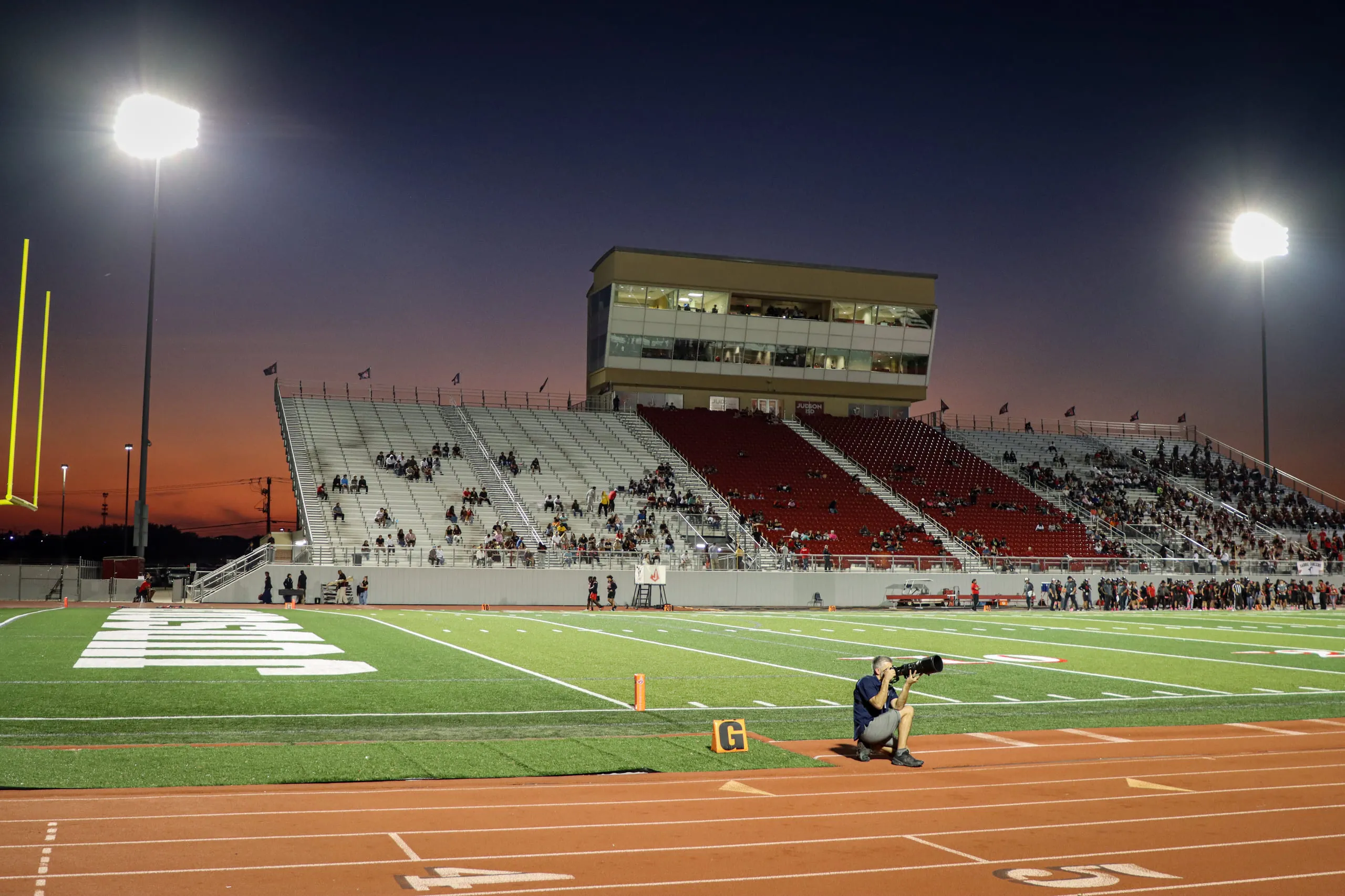 A sideline photographer kneeling with a long lens at a high school football stadium at dusk, floodlights on and the stands filling.