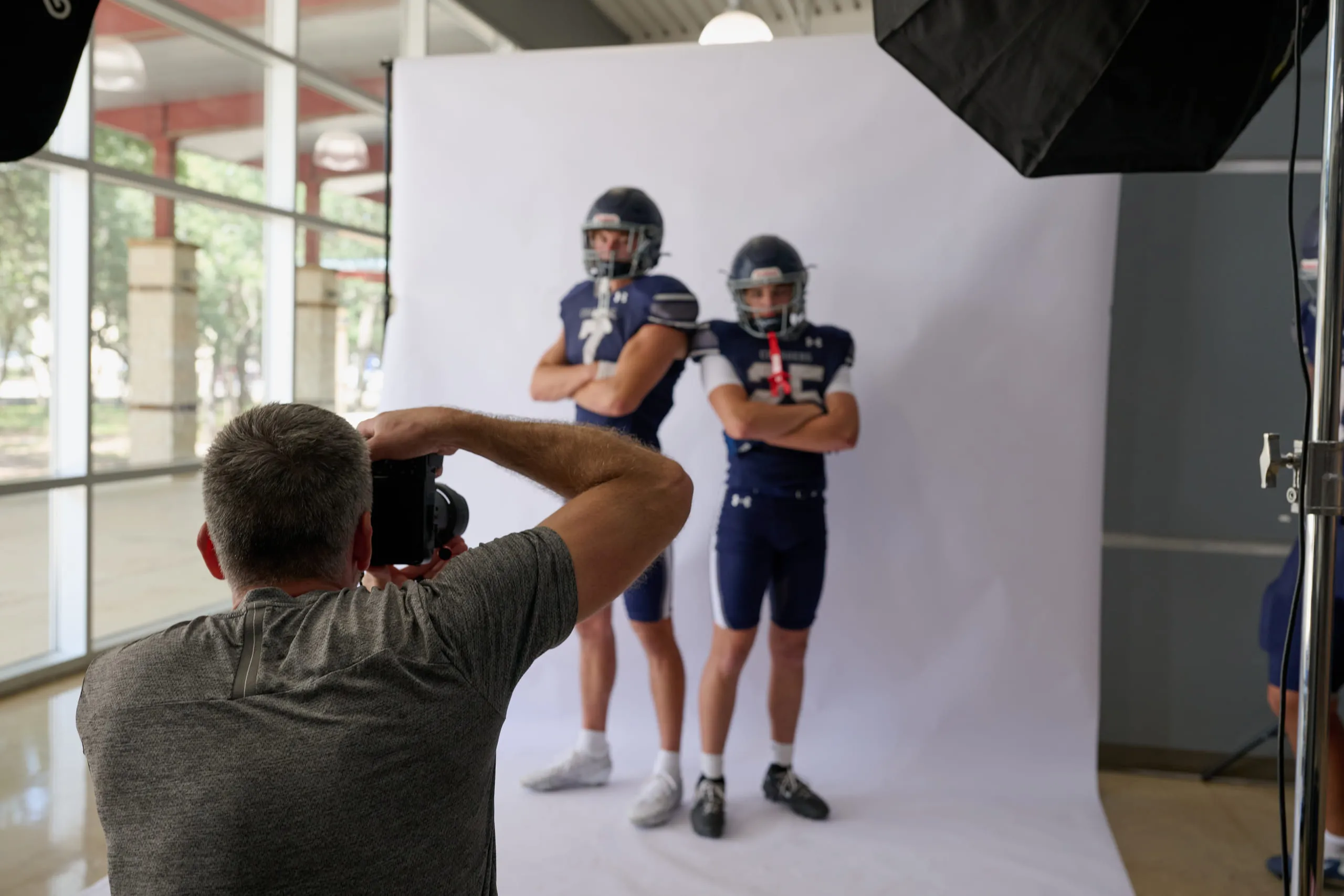 The on-site media-day studio: a photographer shooting two football players in helmets and shoulder pads against a lit white backdrop, softboxes either side.