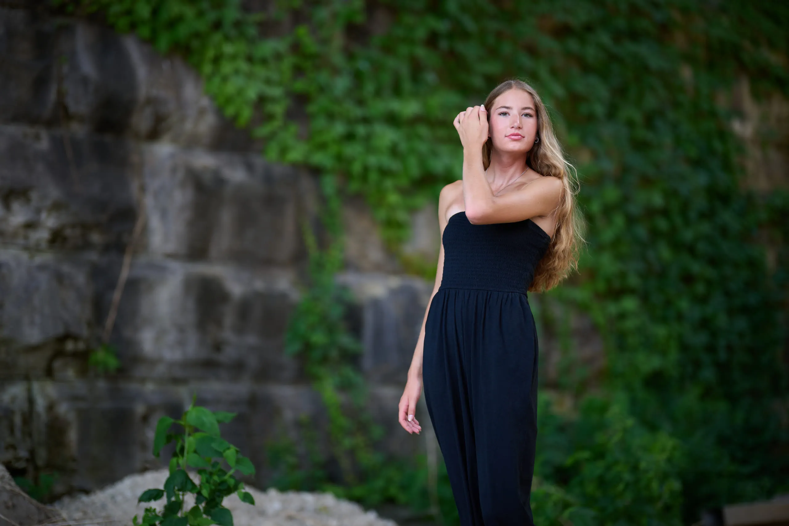 Senior portrait: a young woman in a long black dress beside an ivy-covered stone wall.